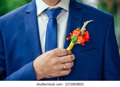 Young groom in a stylish blue wedding suit holds a wedding attribute a boutonniere with his hand with golden ring on his finger. Boutonniere flower roses in bright colors on a groom suit, selective