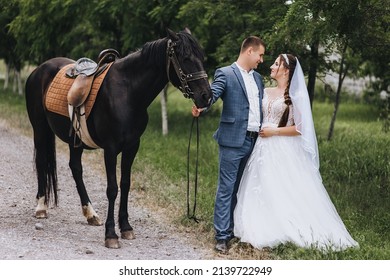 A young groom in a suit and a beautiful bride in a white long dress hug in a village outdoors, walking with a black horse along a country road. Wedding photography, portrait.