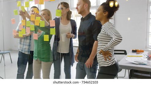 Young Group Of Diverse People Standing Behind Glass Wall Of Office Putting Bright Stickers On It And Collaborating On New Project.