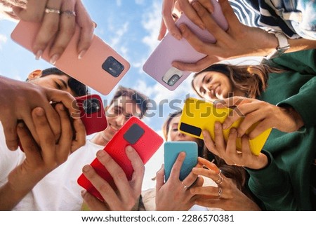 Young group of people using mobile phone device standing together in circle outdoors. Millennial friends addicted to social media app, betting or playing video game on platform online.