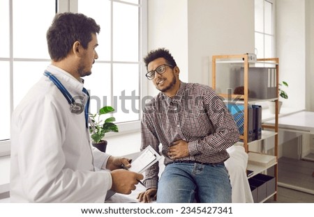 Young guy with acute pain in right side talking to doctor. Adult ethnic patient with stomachache, kidney bloating or cirrhosis asking for help during consultation appointment in exam room at hospital