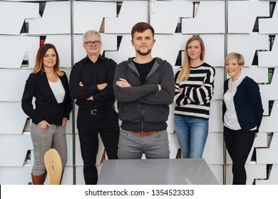 Young Guy With Arms Crossed In Front Of A Small Business Team Standing And Posing To A Group Photo In Their Office. Five Confident People In Casual Clothes Against Decorative White Wall