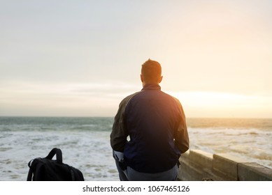 A young guy with a backpack sits by the sea at dawn and admires