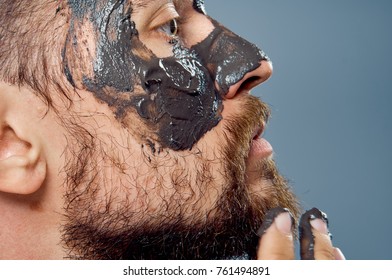 A young guy with a beard on a gray background applies a cosmetic clay mask for the face.