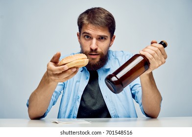 Young guy with a beard on a light background holds a bottle and a hamburger.