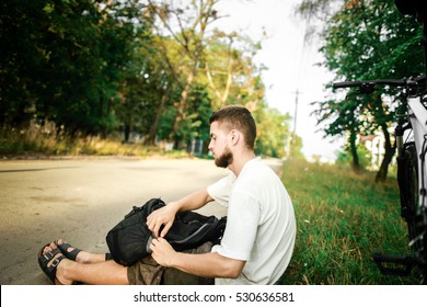 A young guy with a beard trip with a backpack on the white bike, he sitting and relaxing.