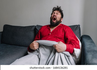 Young Guy With Black Beard Sitting On A Couch And Yawning, Sleepy Man In Red Pajamas Feeling Very Tired, Sleepless Boy At Home