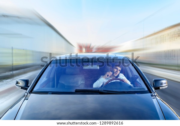 A young guy is a black car driver talking on a\
mobile phone / smartphone, drinking tea, coffee and shaving with an\
electric razor. The concept of not safe driving. Blurred\
background