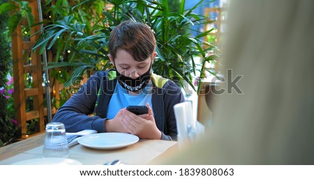 young guy in black designed face mask types on smartphone sitting at restaurant table on terrace and mom hair on foreground