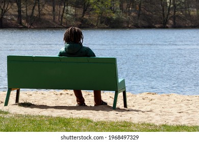 young guy with black dreadlocks sits on a wooden green bench near the lake on a sunny spring day