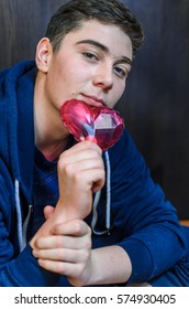 A Young Guy With Blue Eyes, Holding A Pink Glass Heart . Creative Person Looks Through The Prism Of The Heart. The Guy Has A Big Scar On Eyebrow And Adolescent Rash