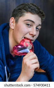 A Young Guy With Blue Eyes, Holding A Pink Glass Heart . Creative Person Looks Through The Prism Of The Heart. The Guy Has A Big Scar On Eyebrow And Adolescent Rash