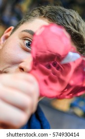 A Young Guy With Blue Eyes, Holding A Pink Glass Heart . Creative Person Looks Through The Prism Of The Heart. The Guy Has A Big Scar On Eyebrow And Adolescent Rash