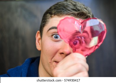 A Young Guy With Blue Eyes, Holding A Pink Glass Heart . Creative Person Looks Through The Prism Of The Heart. The Guy Has A Big Scar On Eyebrow And Adolescent Rash