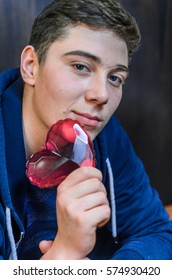 A Young Guy With Blue Eyes, Holding A Pink Glass Heart . Creative Person Looks Through The Prism Of The Heart. The Guy Has A Big Scar On Eyebrow And Adolescent Rash