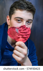 A Young Guy With Blue Eyes, Holding A Pink Glass Heart . Creative Person Looks Through The Prism Of The Heart. The Guy Has A Big Scar On Eyebrow And Adolescent Rash