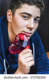 A Young Guy With Blue Eyes, Holding A Pink Glass Heart . Creative Person Looks Through The Prism Of The Heart. The Guy Has A Big Scar On Eyebrow And Adolescent Rash