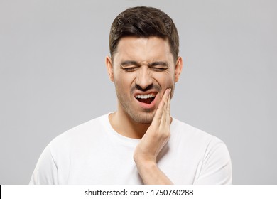 Young Guy With Closed Eyes Suffering From Severe Toothache, Touching Jaw With Fingers Trying To Ease Strong Tooth Pain, Isolated On Gray Background