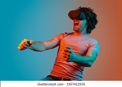 Young Guy With Curly Hair Screaming And Driving Virtual Car While Playing VR Racing Game Under Blue And Orange Light 