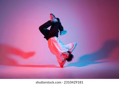 young guy dancer is dancing break in neon lighting, male acrobat is doing trick and dance exercise on red blue background, hip hop performer is moving and standing on his hands