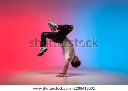 young guy dancer is dancing break in neon lighting, male acrobat is doing trick and dance exercise on red blue background, hip hop performer is moving and standing on his hands