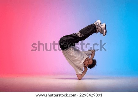 young guy dancer is dancing break in neon lighting, male acrobat is doing trick and dance exercise on red blue background, hip hop performer is moving and standing on his hands