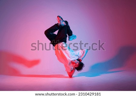 young guy dancer is dancing break in neon lighting, male acrobat is doing trick and dance exercise on red blue background, hip hop performer is moving and standing on his hands