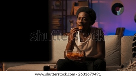 A young guy with distinctive features and an afro sits upright on a sofa, watching a TV show alone in the living room, eating chips and reacting with joy to thrilling scenes on screen.