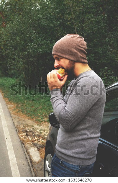 Young guy eating\
a burger near a car on an empty road. Food on the trip. Food on the\
go. Autumn travel. Fast\
food