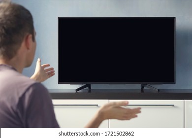 A Young Guy Emotionally Watching TV And Waving His Hands In Discontent. Blank Screen For Design. Frustrated Man Sitting On Sofa In Front Television With No Signal.