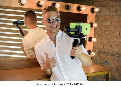 Young guy filming a blog on a mobile device using an electronic image stabilizer at the studio against the background of a mirror with illumination. Blond European man in sunglasses shoots a video