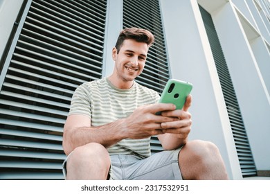 Young guy with friendly expression smiling and having fun playing a video game with his cellphone sitting outdoors. Caucasian teenage stundent enjoying using a mobile phone for watch the social media - Powered by Shutterstock