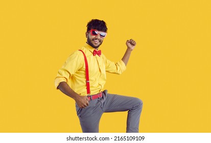 Young guy in funny party outfit having fun in studio. Joyful Indian man with trendy haircut wearing yellow shirt, pants with suspenders, red bowtie and sunglasses celebrating happy event and dancing