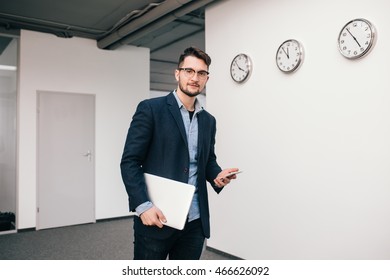 Young Guy In Glasses Is  Walking In Office. He Wears Blue Shirt, Dark Jacket, Jeans And Beard. He Holds  Phone And Laptop In Hands. Looking To The Camera.