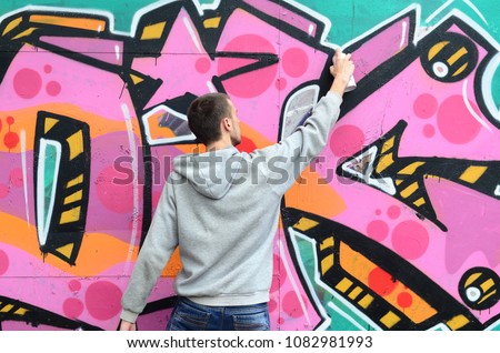 A young guy in a gray hoodie paints graffiti in pink and green colors on a wall in rainy weather