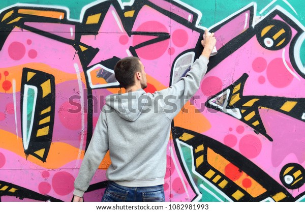 A young guy in a gray hoodie\
paints graffiti in pink and green colors on a wall in rainy\
weather