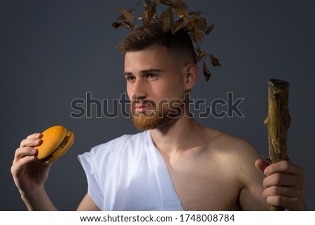 a young guy in the guise of an Olympic god in a laurel vignette with a staff, holds a burgur in his hand, looking at him. Studio photo on a gray background.