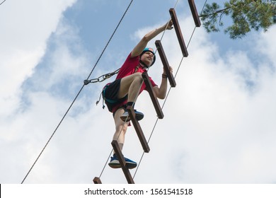 A Young Guy In A Helmet Climbs Up A Rope Ladder Against The Blue Sky. Mountaineering And Climbing Wall For Tourists And Vacationers. Sports Lifestyle.