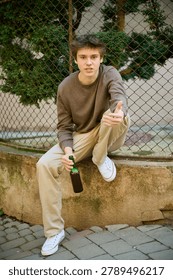 Young guy holds bottle while crouching in urban setting and gestures ahead. Serious teenager by street fence shows inner conflict and vulnerability. Concept of alcohol abuse among minors