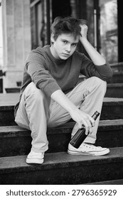 Young guy holds bottle while sitting on city stairs. Troubled teenager in casual clothes appears lonely and lost in thought. Concept of youth drinking problem and vulnerability