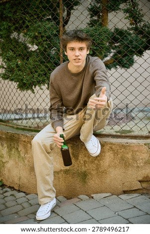 Young guy holds bottle while crouching in urban setting and gestures ahead. Serious teenager by street fence shows inner conflict and vulnerability. Concept of alcohol abuse among minors