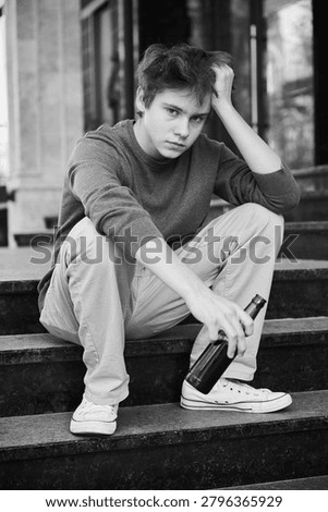 Young guy holds bottle while sitting on city stairs. Troubled teenager in casual clothes appears lonely and lost in thought. Concept of youth drinking problem and vulnerability