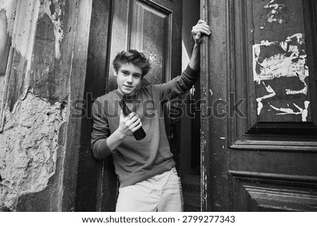 Young guy holds bottle while stepping through old door. Troubled teenager in neglected city setting appears tense and isolated. Concept of youth drinking problem and vulnerability