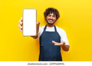 young guy Indian waiter in apron shows blank screen of smartphone on yellow isolated background, male Indian barista in uniform advertises mobile phone online - Powered by Shutterstock