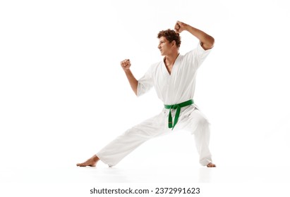 Young guy, karate sportsman in white kimono and green belt standing in pose to fight isolated on white studio background. Concept of martial arts, combat sport, energy, strength, health. Ad
