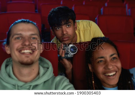 Young guy looking focused, using his video camera for recording a pirated movie at the cinema. Selective focus. Horizontal shot