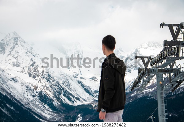 A young guy looks at\
the snowy mountains from above. Nice view of the snowy mountains\
and the cable car.