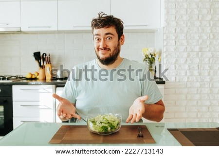 A young guy, a man, sits at a table, looks at the camera and pretends that he really wants to eat this salad. He was tired of dieting and following the principles of healthy eating. Stop diet.