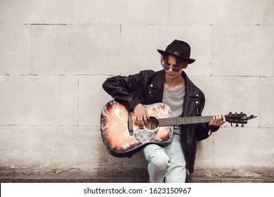 Young guy musician wearing hat and sunglasses standing leaning back on wall playing guitar concentrated - Shutterstock ID 1623150967