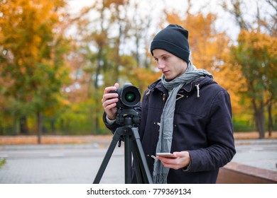 A young guy in park sets up a camera and holds a mobile phone in his hand. Outdoors.